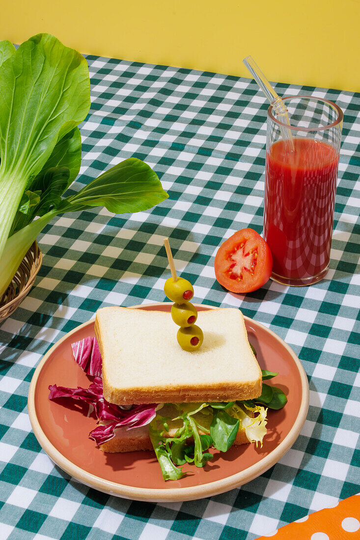 Appetitliches gesundes Sandwich mit frischem Salat auf einem Teller mit Oliven neben einem Glas Tomatensaft mit Glasstrohhalm auf einem karierten Tischtuch