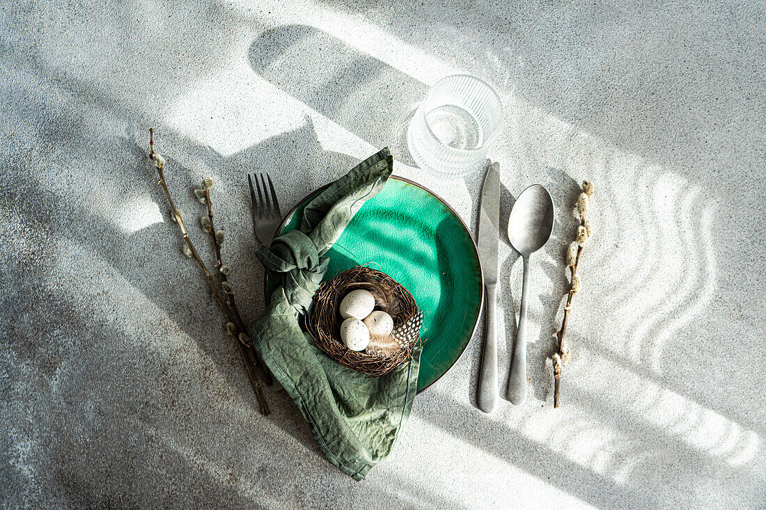 Top view of Easter table setting, showcasing a vibrant green ceramic plate with a small nest with speckled eggs and feathers, placed on gray surface with napkin, cutlery and glass of water