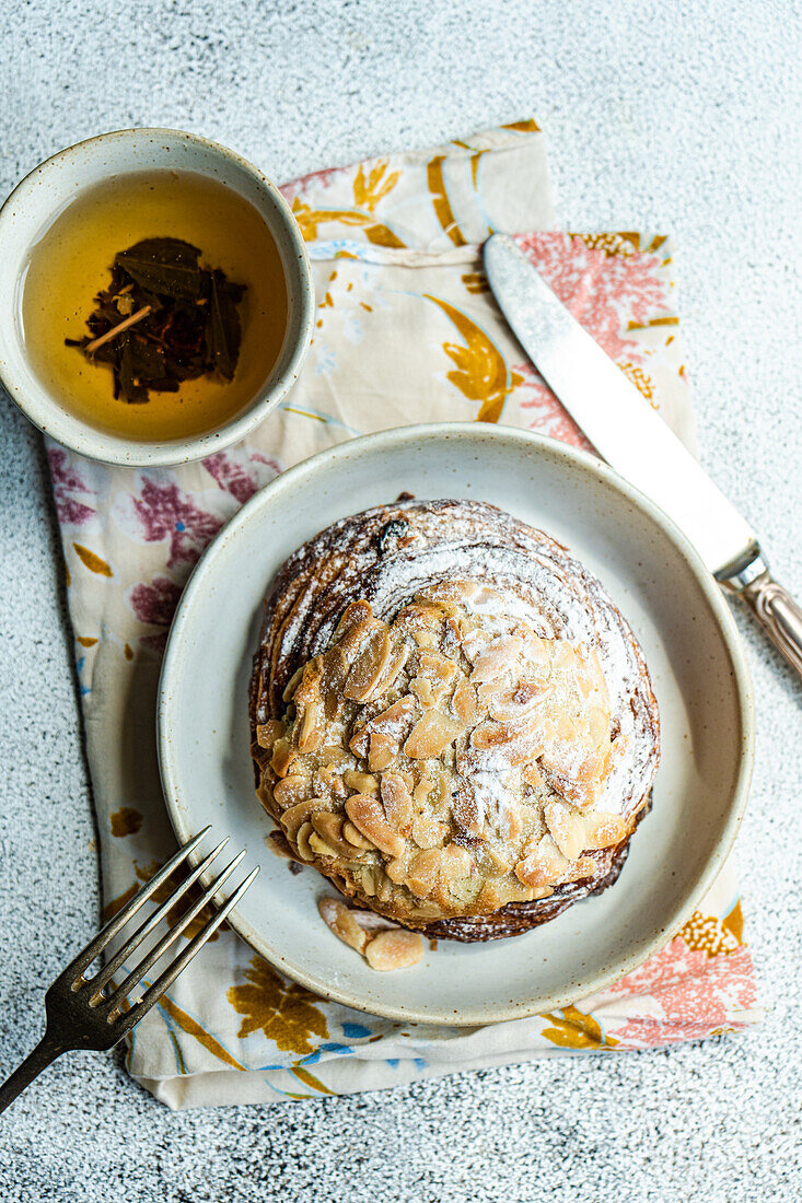 Top view of sweet almond pastry on napkin with fork and knife with cup of green tea against gray background in sunlight