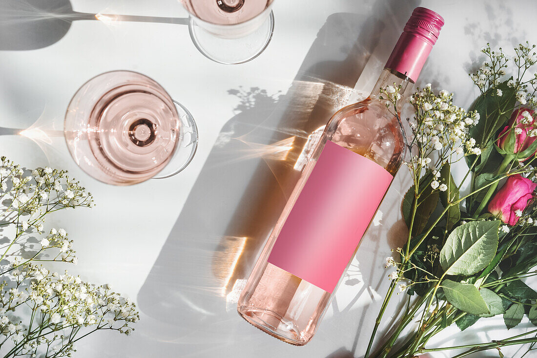 Rose Weinflasche mit leeren rosa Mock-up-Etikett, zwei Weingläser und romantischen Blumenstrauß auf weißem Hintergrund mit Sonnenlicht und Schatten. Modernes Wein-Konzept. Ansicht von oben