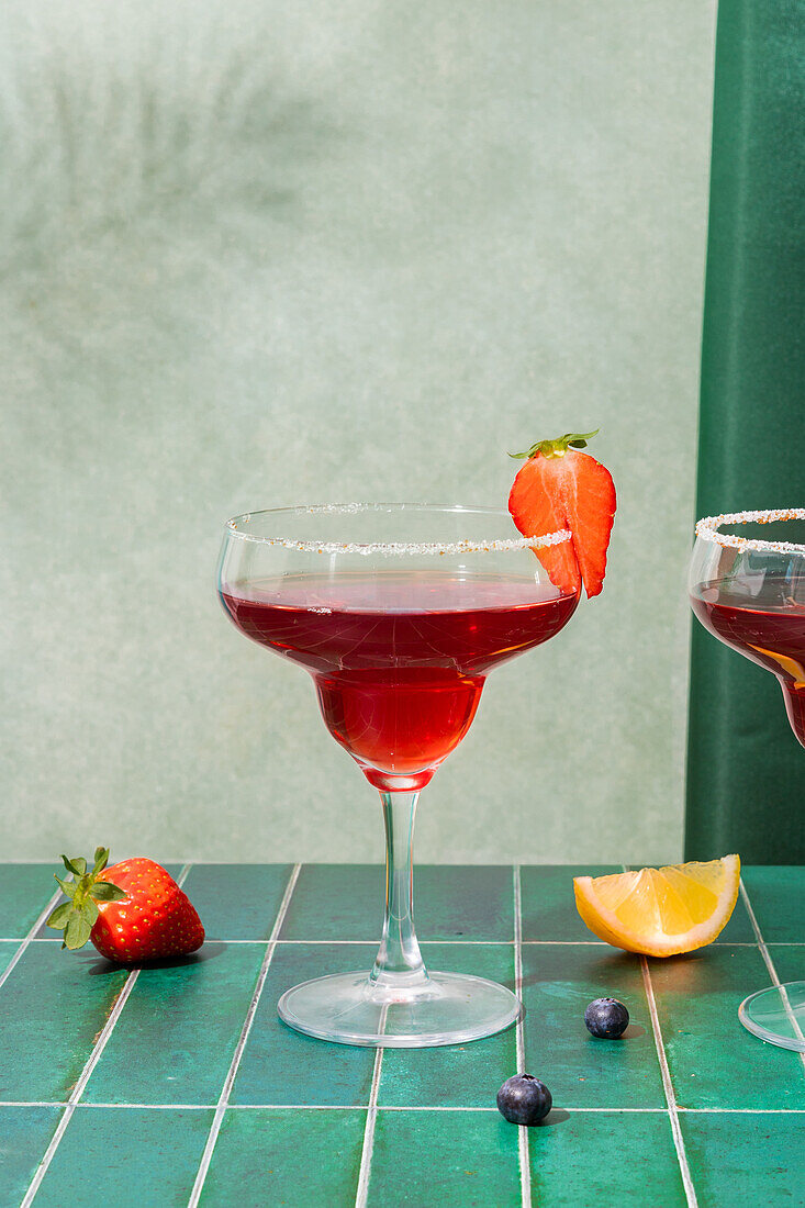 Gläser mit erfrischenden roten alkoholischen Cocktails mit Erdbeeren und Strohhalm, serviert auf einem Tablett mit Beeren und Zitronenscheibe auf dem Tisch