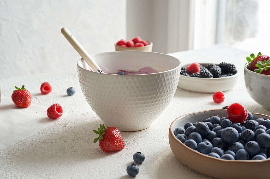 Schale mit Naturjoghurt und einigen frischen Früchten darin: Himbeeren, Blaubeeren, Erdbeeren und Brombeeren neben Tellern mit frischen Früchten im Tageslicht