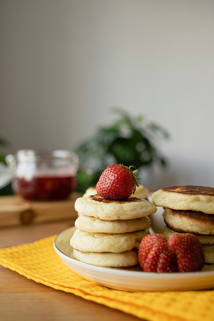 Delicious sweet strawberries placed over stacked pancake on plate over tablecloth on wooden table with strawberry jam in bright room