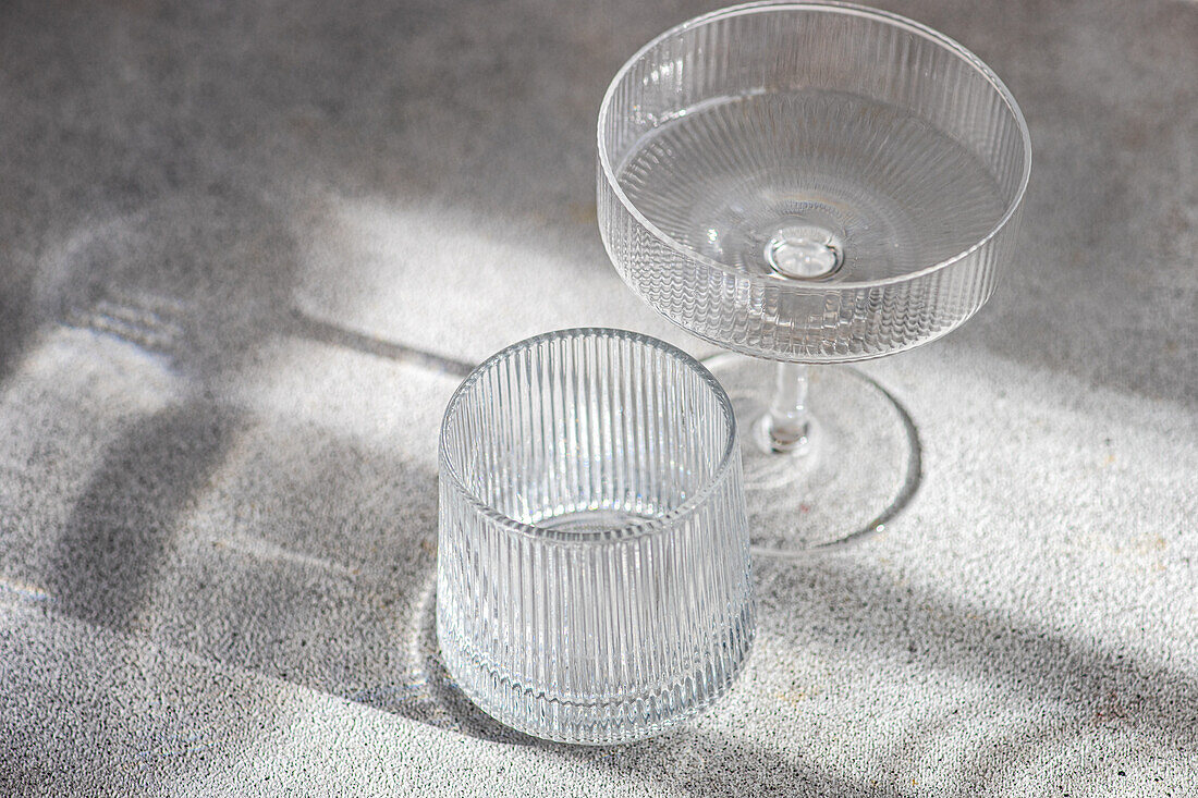 A crystal clear cocktail glass and tumbler sit on a textured surface, their distinct shadows cast by soft, natural light