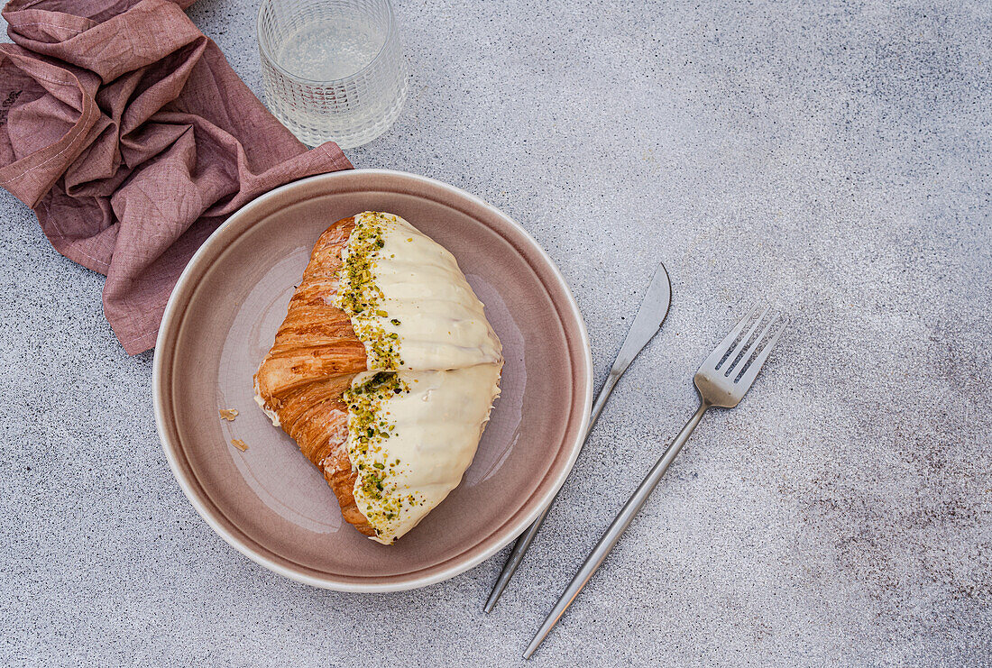 From above golden croissant partially covered with white icing and pistachio pieces, on a round beige plate beside a knife and fork, set on a grey background with a folded brown napkin