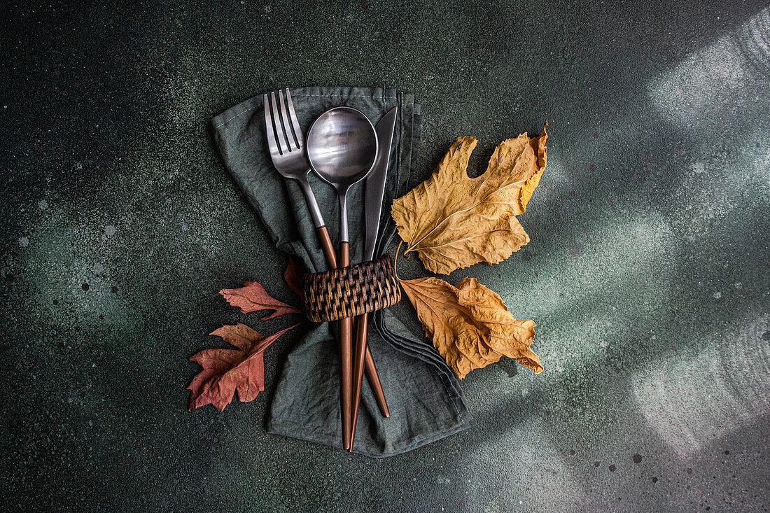 Top view of autumnal table setting with napkin, knife, spoon and fork place on gray surface near colorful leaves