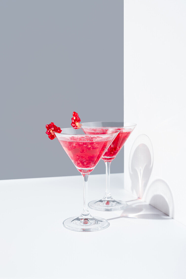 Glasses filled with red pomegranate cocktails served with pomegranate seeds against a gray and white backdrop, casting shadows