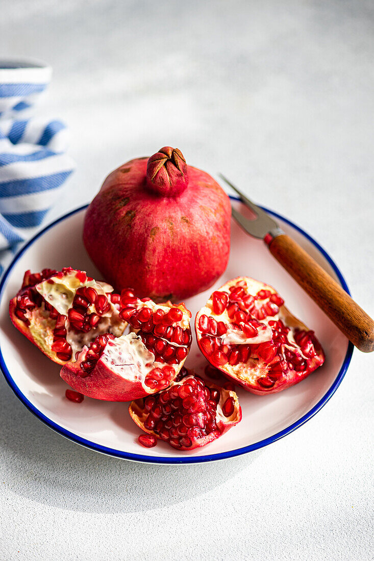 A ripe pomegranate open on a blue-trimmed plate, revealing its red seeds, accompanied by a wooden handled knife..