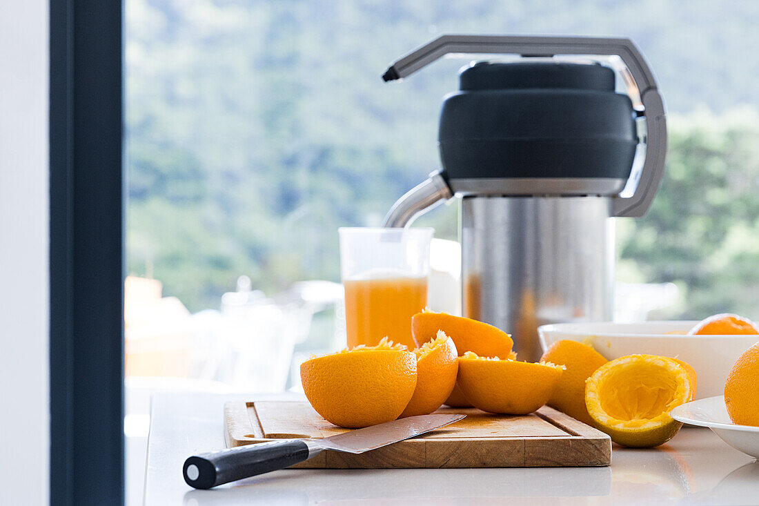 Glas mit frischem Orangensaft und Saftpresse auf dem Küchentisch in der Nähe von extrahierten Orangenscheiben, die mit einem scharfen Messer auf einem Schneidebrett im Tageslicht vor einem unscharfen Hintergrund platziert sind
