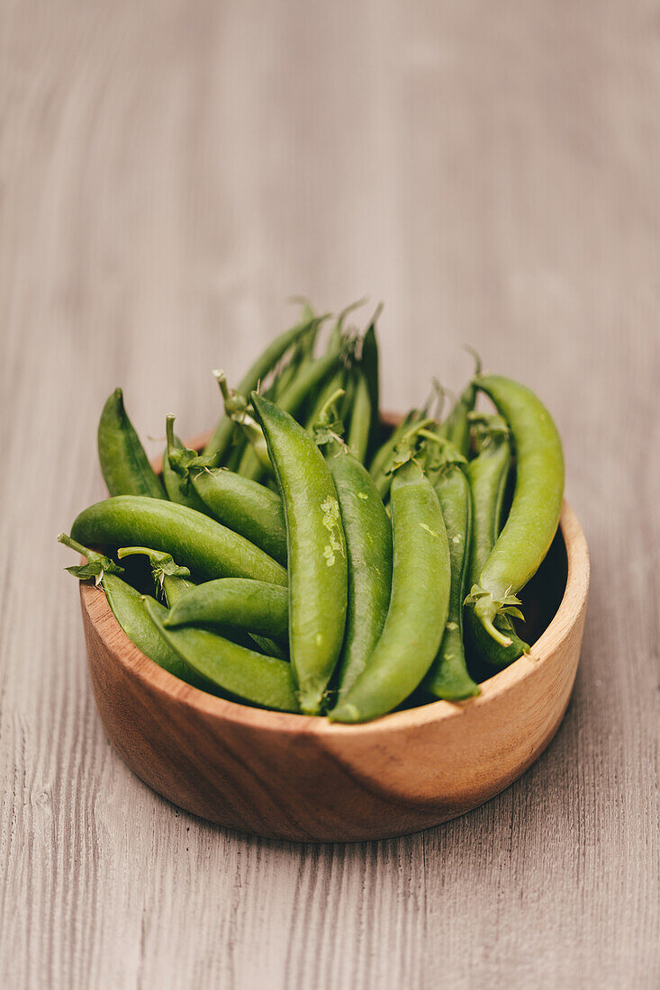 A wooden bowl filled with ripe green peas is set against a textured wooden backdrop, presenting a natural and rustic image.