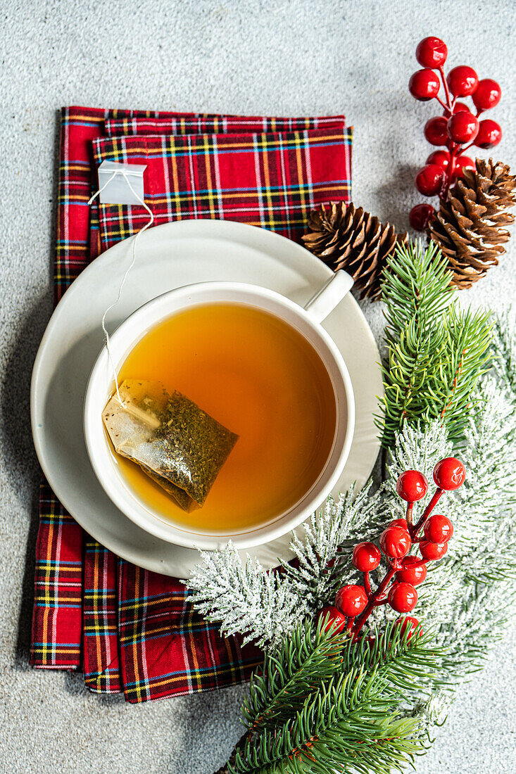 Top view of Christmas cup of tea placed on red napkin near holly and pine cones on gray table