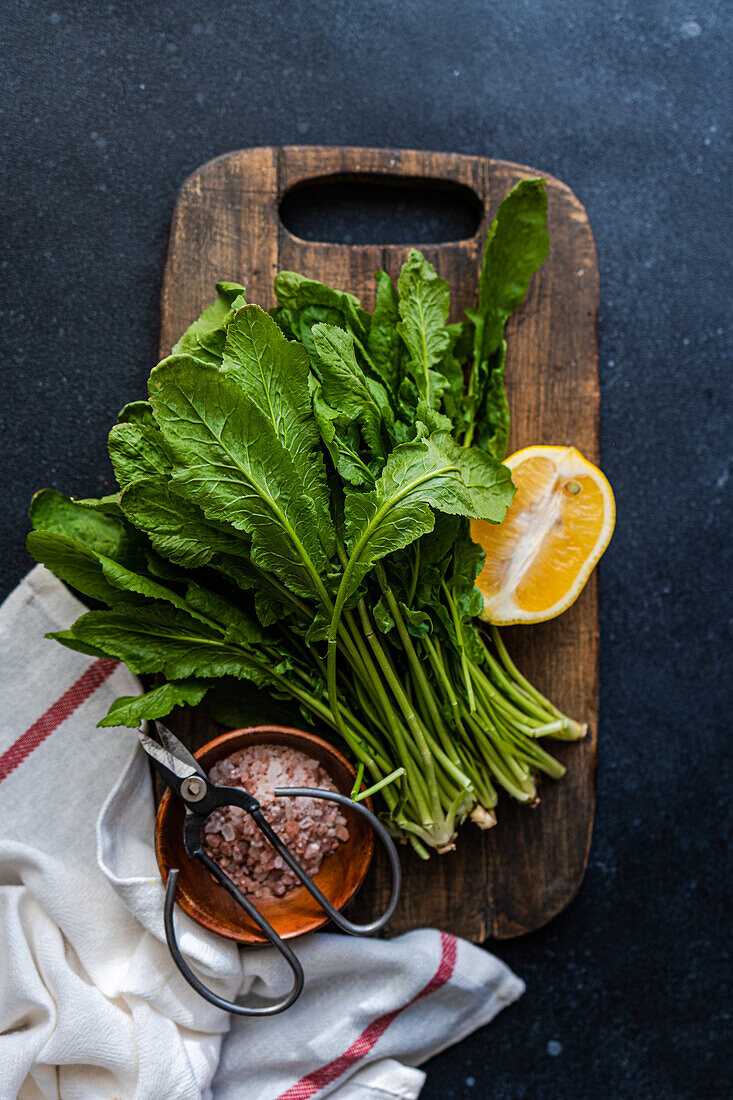 From above of fresh arugula leaves with a slice of lemon and a bowl of salt on a wooden cutting board, accented by kitchen scissors and a striped towel