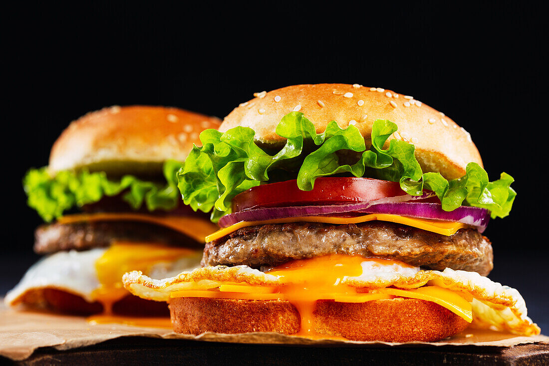 Delicious burgers with fresh buns and vegetables served on yummy patties ans cheese against black background on parchment paper