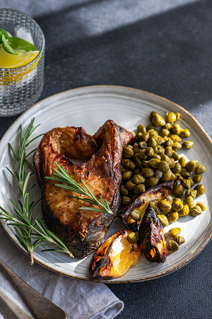 From above grilled salmon steak served with fermented capers and a wedge of lemon garnished with fresh rosemary on a rustic ceramic plate with vintage cutlery