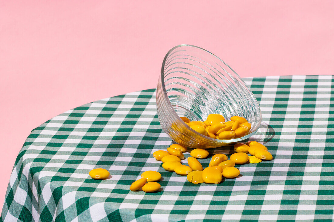Transparent glass bowl with yellow lupin beans placed on checkered table against pink background