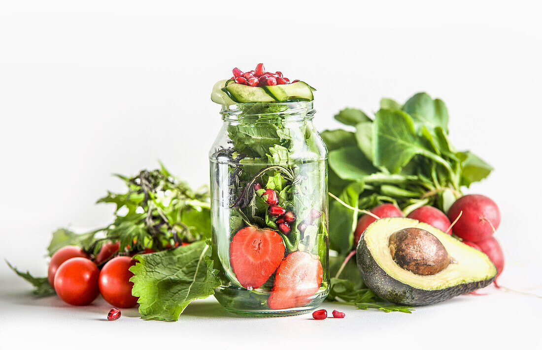 Grüner Salat im Glas mit Zutaten: grüne Salatblätter, Tomate, Erdbeere, Avocado, Gurke und Granatapfelkerne auf weißem Hintergrund. Gesundes Essen zum Mitnehmen mit Gemüse und Obst. Frontansicht