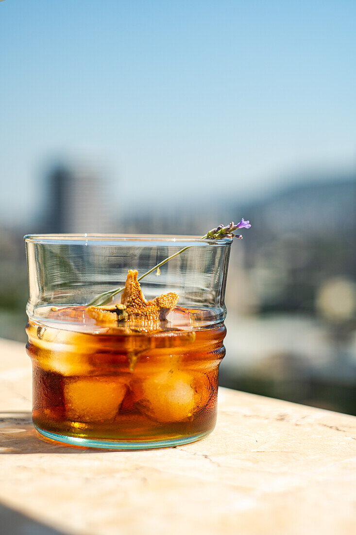 Glas Whiskey mit Eis, Orangenschale und Blume auf weißer Fläche vor unscharfem Gebäude