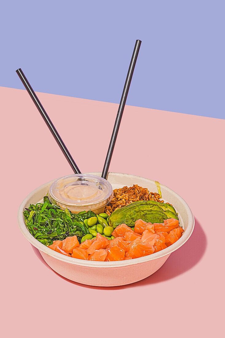 High angle of poke bowl filled with fresh salmon, green edamame beans, crunchy seaweed salad, creamy avocado, and crunchy granola, accompanied by a light dipping sauce and chopsticks, against a dual-toned pastel background