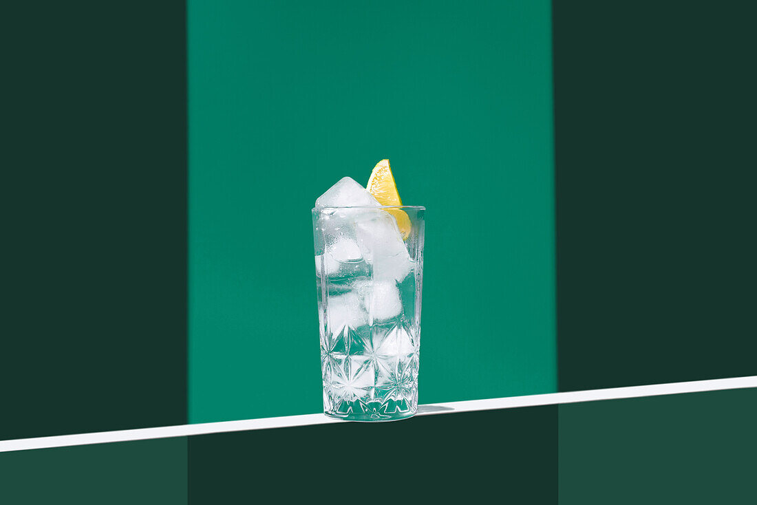 Eine minimalistische Szene mit einem transparenten Becher, gefüllt mit Gin Tonic und einer Limettenscheibe, vor einem zweifarbigen grünen Hintergrund