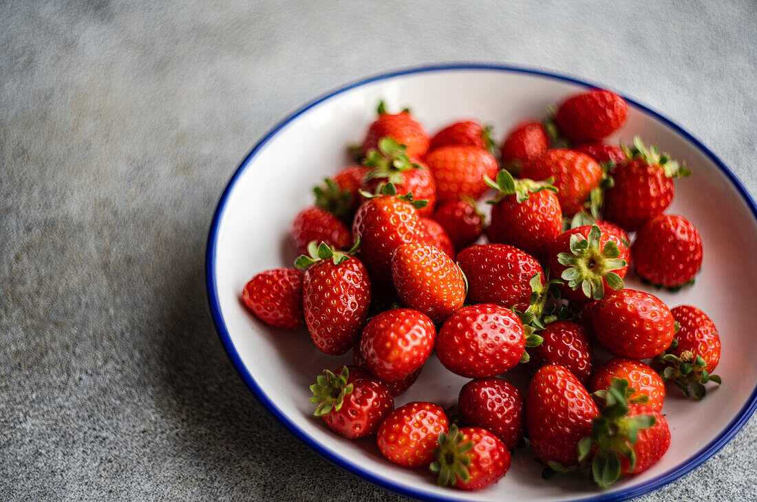 Close-up of vibrant organic strawberries in a white dish with a blue trim, set on a muted gray background.