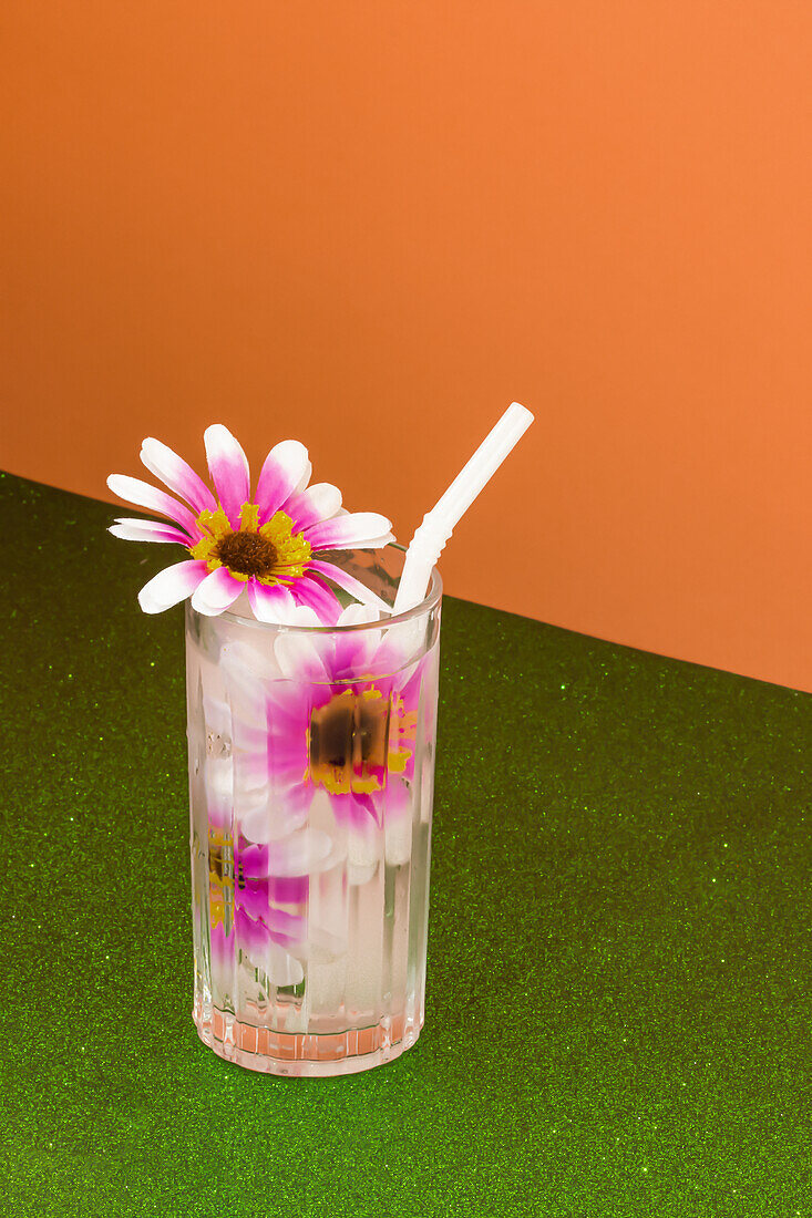 Transparentes Glas mit erfrischendem Kaltgetränk, verziert mit rosa Blumen und Strohhalm, auf grüner Fläche vor leuchtend oranger Wand
