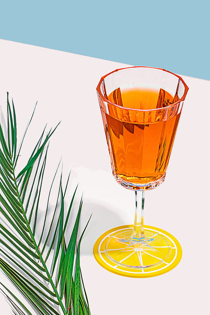 Ein stilvoller bernsteinfarbener Cocktail in einem Kristallglas, der auf einem Zitrusuntersetzer neben einem Palmwedel vor einem pastellfarbenen Hintergrund ruht