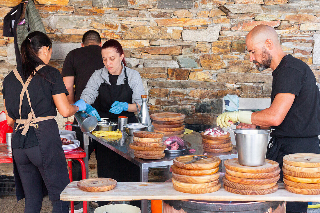 Ein Team von Köchen, gekleidet in schwarzer Schürze und blauen Handschuhen, bereitet fleißig Gerichte mit Tintenfisch auf dem Tisch vor einem rustikalen Steinmauer-Hintergrund zu