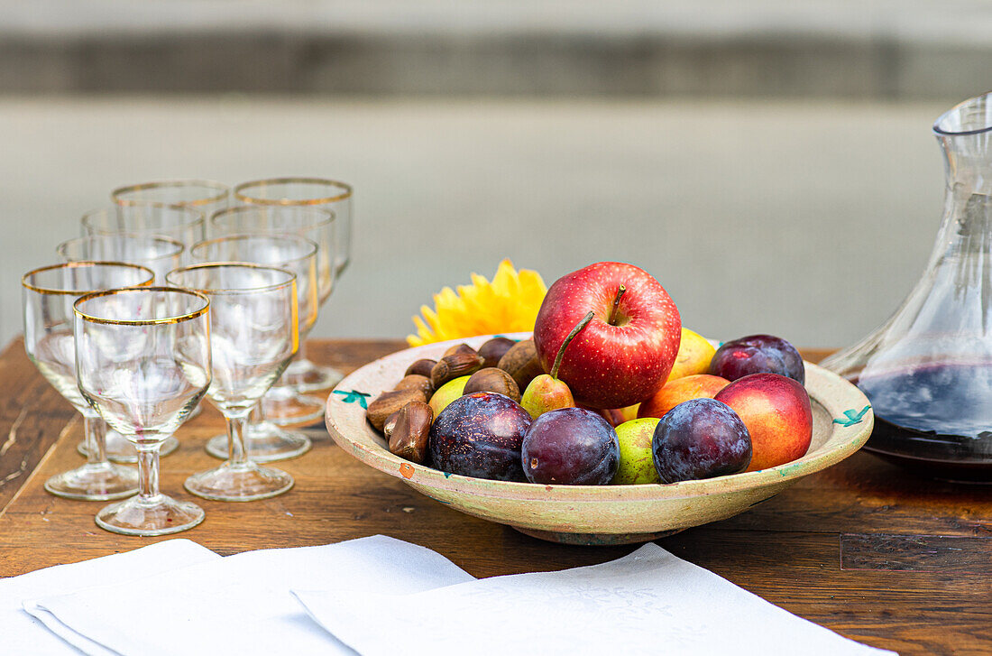 A wooden table displaying an array of fresh fruits in a bowl, accompanied by empty wine glasses and a pitcher of juice, set against a muted backdrop.