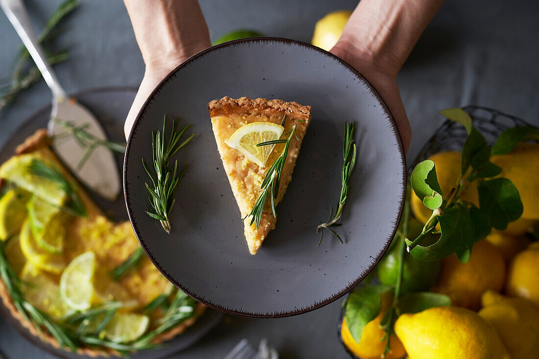 From above anonymous person holding a plate with a slice of lemon pie with decorated with lemon slice and rosemary sprig on table in the kitchen