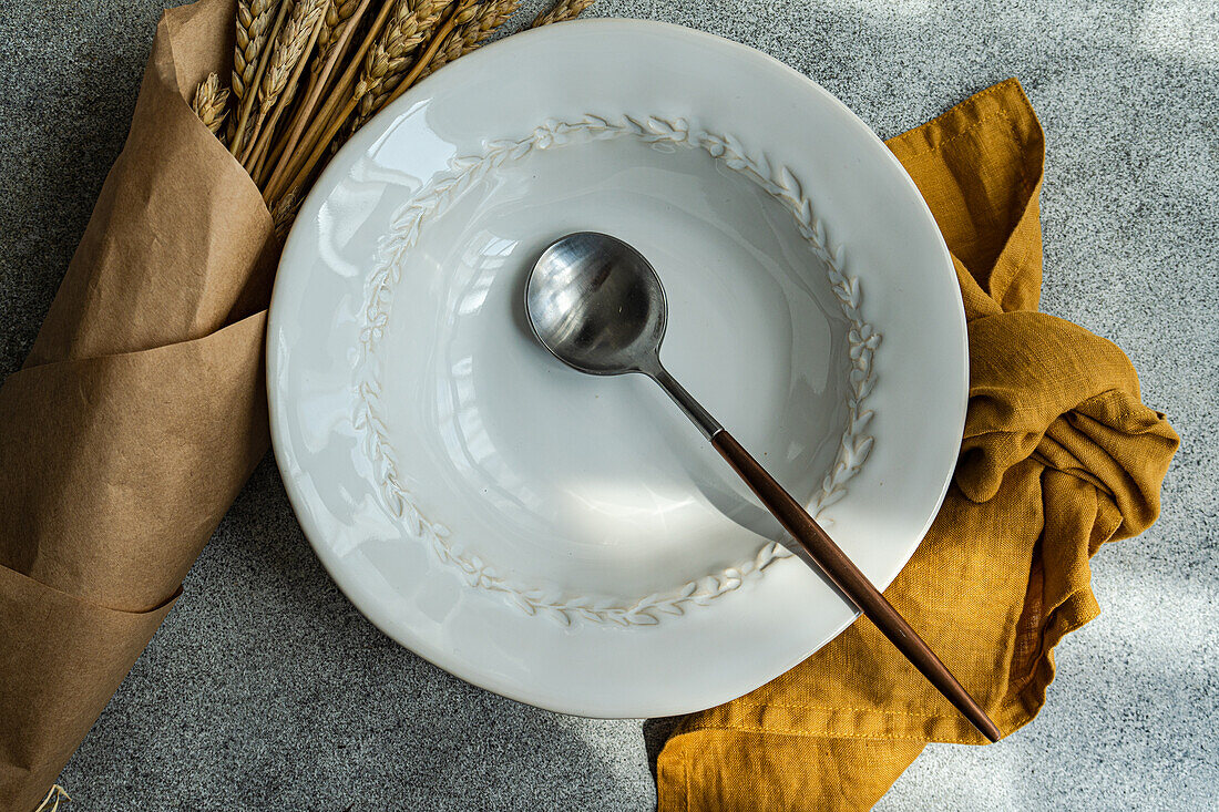 Draufsicht auf einen gedeckten Sommertisch mit Teller und Löffel neben Serviette und Weizen auf einem grauen Tisch