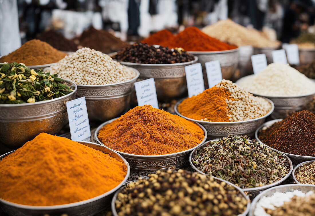 Reihen von gemischten farbigen pulverisierten Gewürzen und Samen in Schalen an einem lokalen Marktstand vor einem unscharfen, geschäftigen Markthintergrund