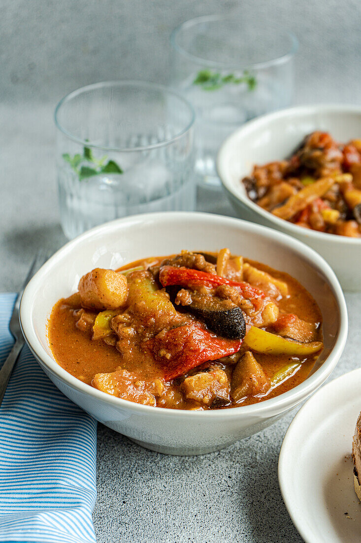 High angle of traditional seasonal Georgian dish - Adjapsandali with stewed vegetables like potato, bell peppers, tomato, egg plant, coriander and Georgian spices placed on gray table near napkin and glass with water