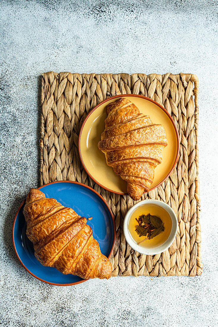 Draufsicht auf frisch gebackene Croissants auf bunten Keramiktellern auf brauner Serviette neben einer Tasse grünem Tee vor grauem Hintergrund
