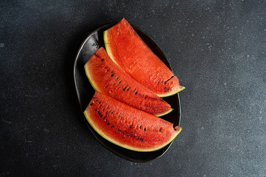 Draufsicht auf Scheiben von Wassermelone, serviert auf schwarzem Teller vor dunkler Oberfläche
