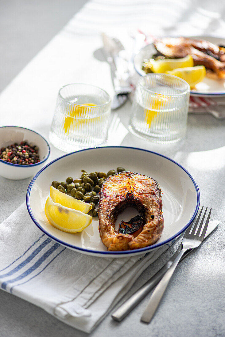 High angle of well-prepared grilled trout steak with capers and lemon served on a white plate with a blue rim placed on striped napkin, cutlery, and glasses of water