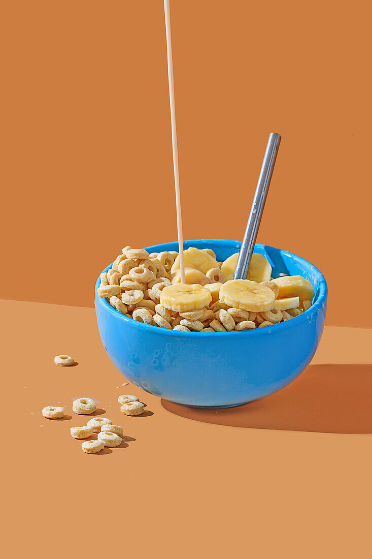 Ein Strom von Milch, der in eine blaue Müslischale mit Cornflakes und Bananenscheiben vor einem orangefarbenen Hintergrund gegossen wird