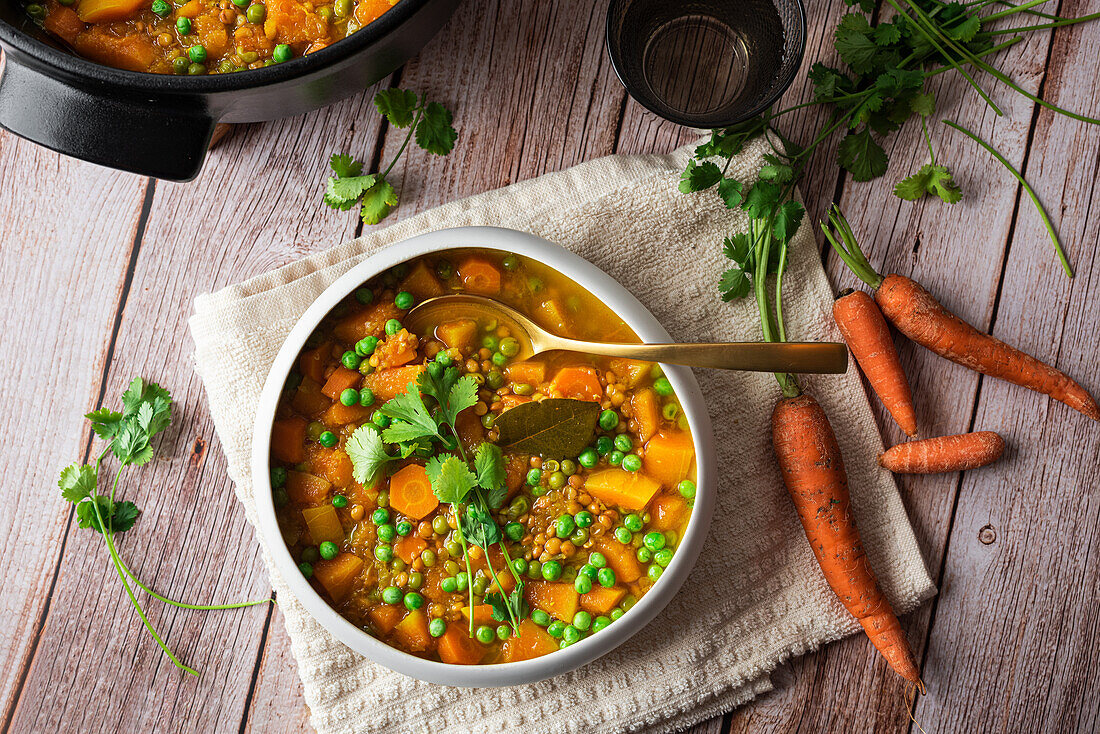 Top view of appetizing fresh cooked soup with carrot and green peas served in white ceramic bowl placed on wooden table and decorated with parsley branch