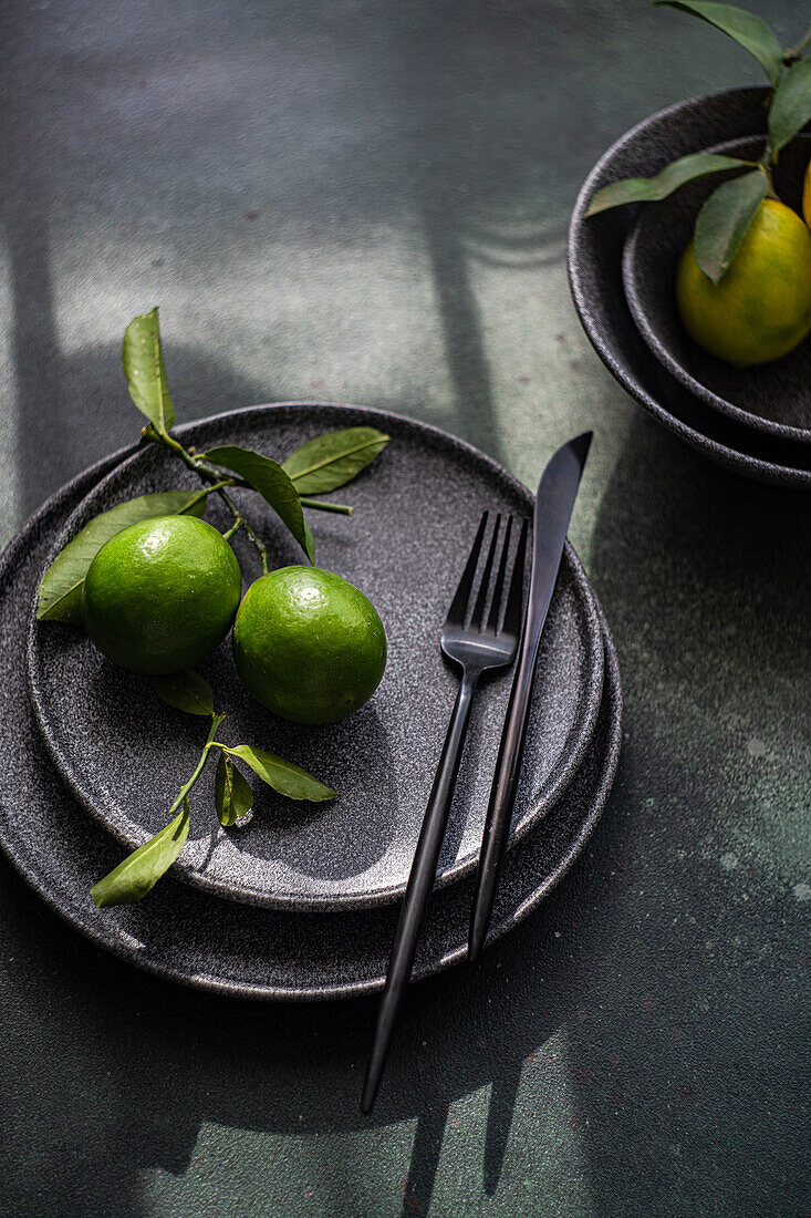 A sophisticated table setting featuring dark plates, cutlery, and fresh green limes on a moody, shadowed surface.