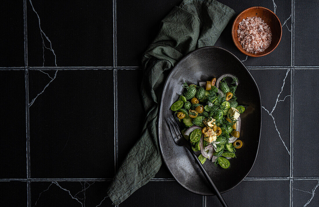 Draufsicht auf einen Gurkensalat mit roten Zwiebeln, Oliven und würzigem Käse, serviert auf einem schwarzen Teller mit Gabel neben einer Serviette auf einem dunklen Tisch mit weißen Linien