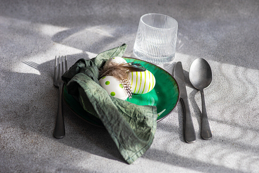 High angle of Easter table setting, showcasing a vibrant green ceramic plate with two decorative Easter eggs adorned with white and green patterns and delicate feathers, placed on gray surface between napkin and cutlery and glass of water