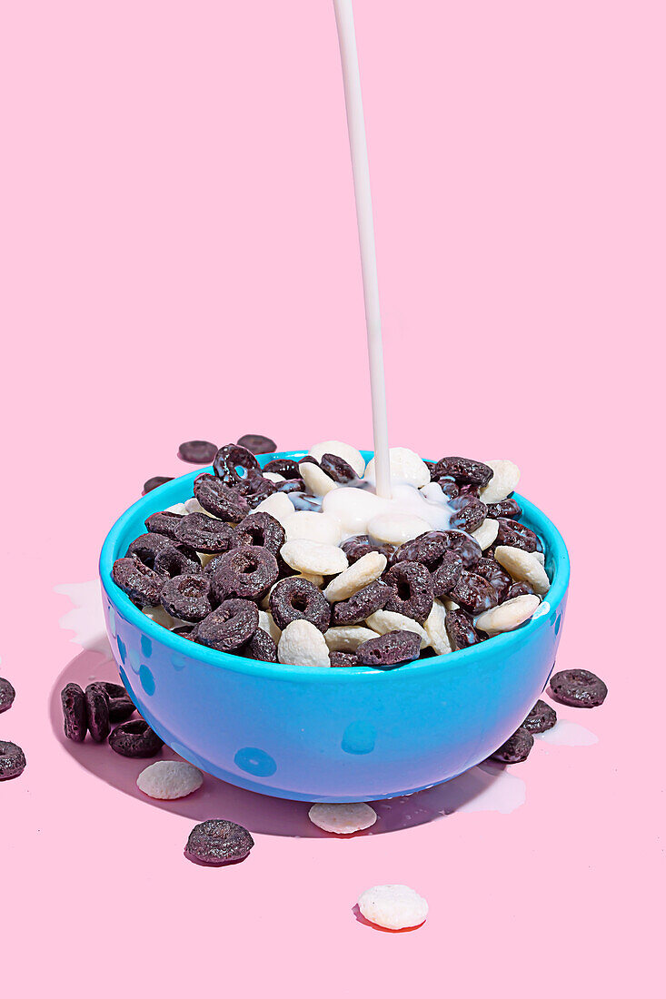 A stream of milk is being poured into a bright blue bowl filled with chocolate-flavored cereal on a pink background.