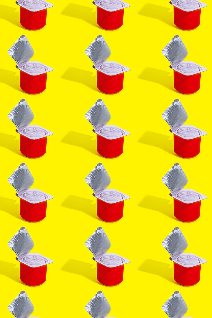 A repetitive pattern of red cups of strawberry yogurt, each with its foil lid opened, arranged neatly on a bright yellow background