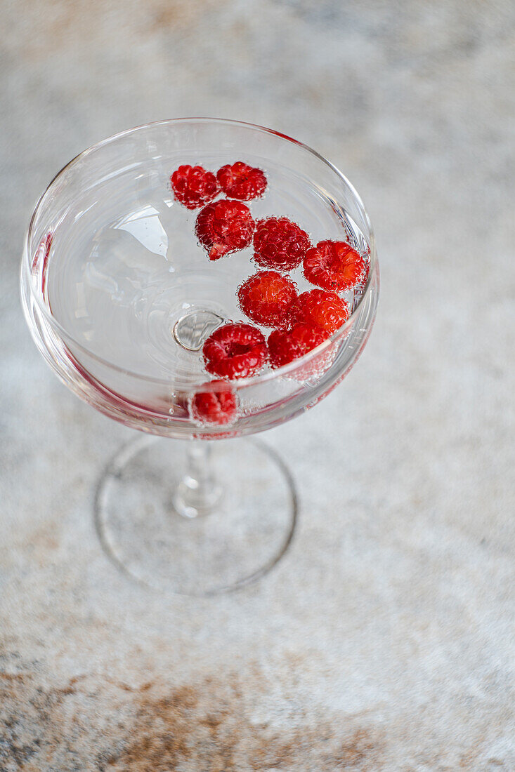 Hoher Winkel eines transparenten, glatten Glasbechers, gefüllt mit erfrischendem Cocktailsekt mit reifen, frischen Himbeeren, der auf einer Tischdecke auf einem Marmortisch steht, vor einem verschwommenen Hintergrund