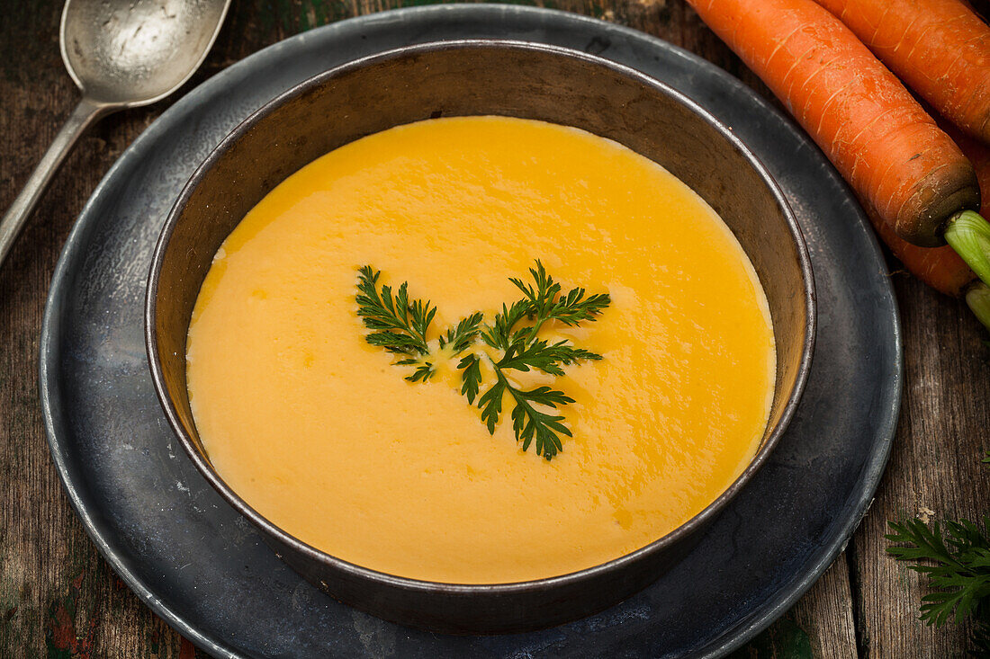 A bowl of vibrant, creamy carrot soup garnished with parsley, served in a rustic setting with fresh carrots and a vintage spoon.
