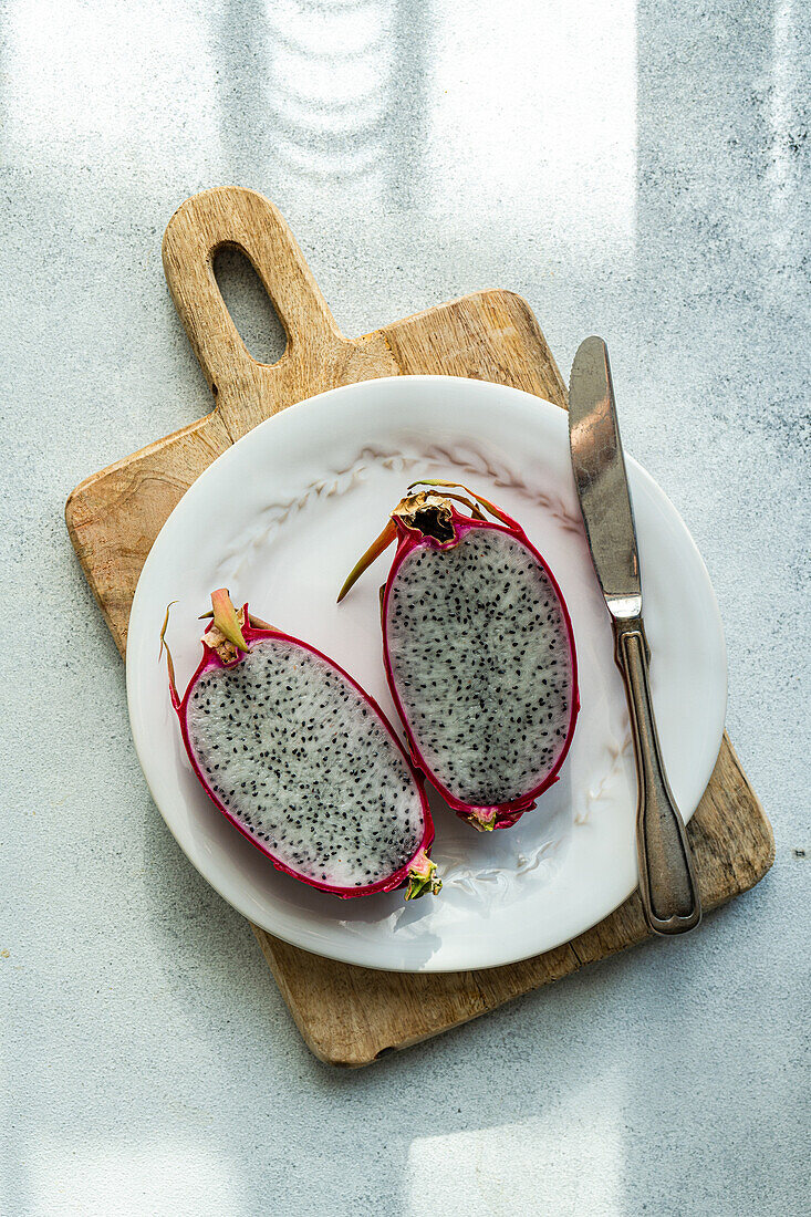 A halved dragonfruit with vibrant pink skin and speckled flesh presented on a wooden serving board with a knife.