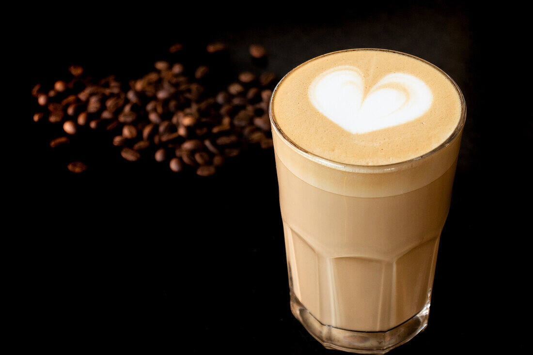 Ein Glas mit aromatischem, heißem Milchkaffee, verziert mit Latte Art, steht auf einem Tisch neben Kaffeebohnen
