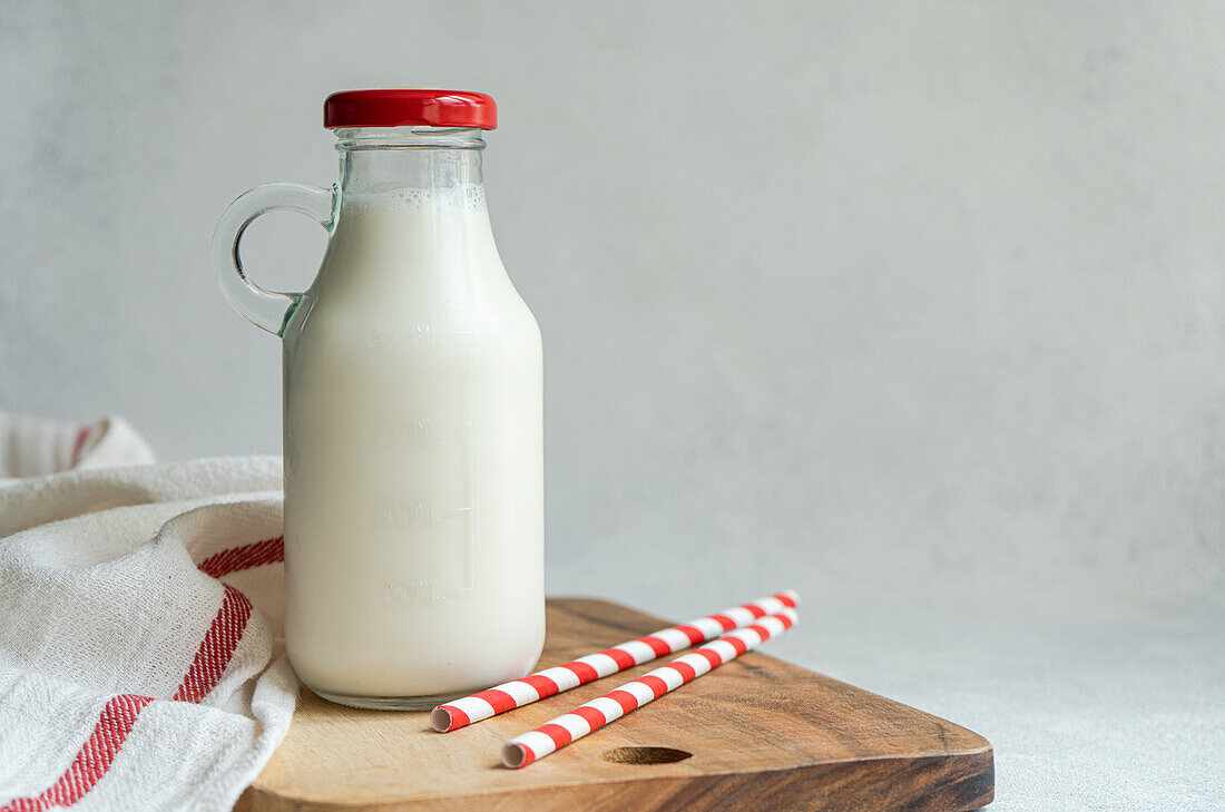 Raw cow milk in vintage closed bottle on cutting board near drinking straws and napkin against gray surface