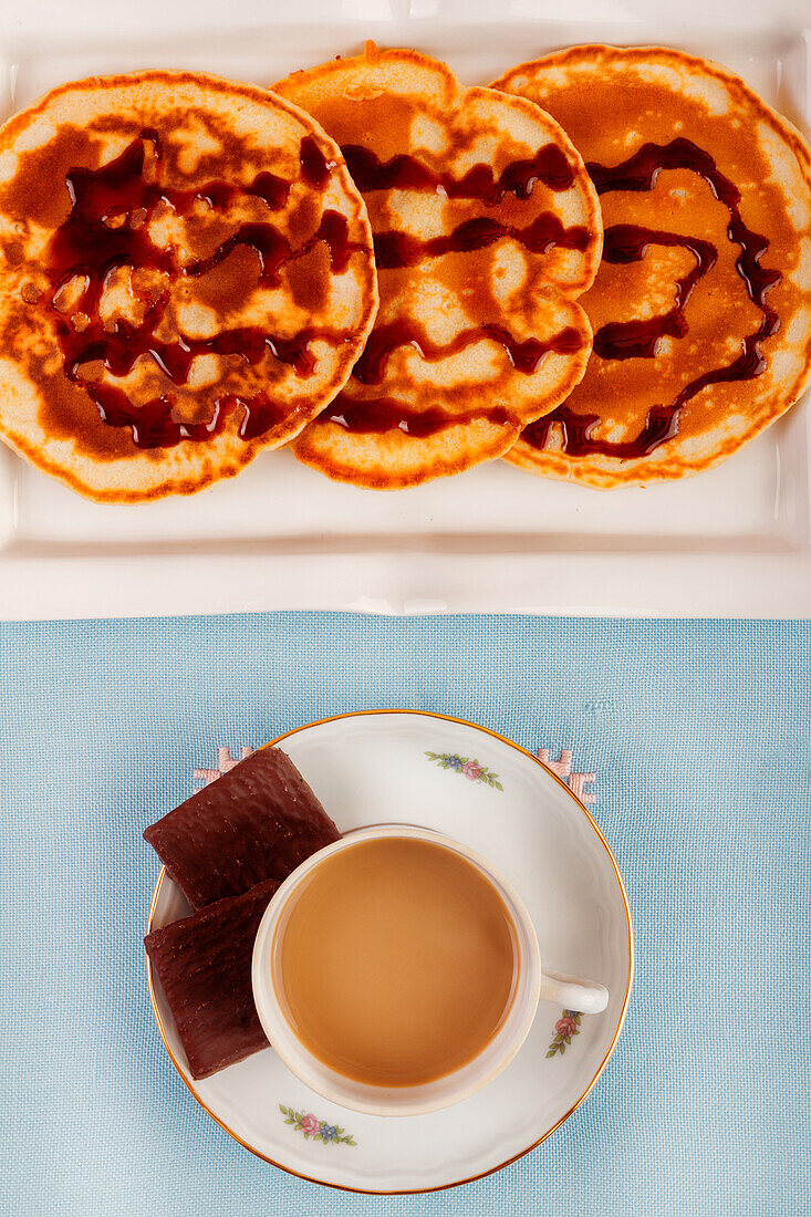 An overhead view of a breakfast scene featuring a cup of coffee with milk and stacked waffles drizzled with syrup on a blue background.