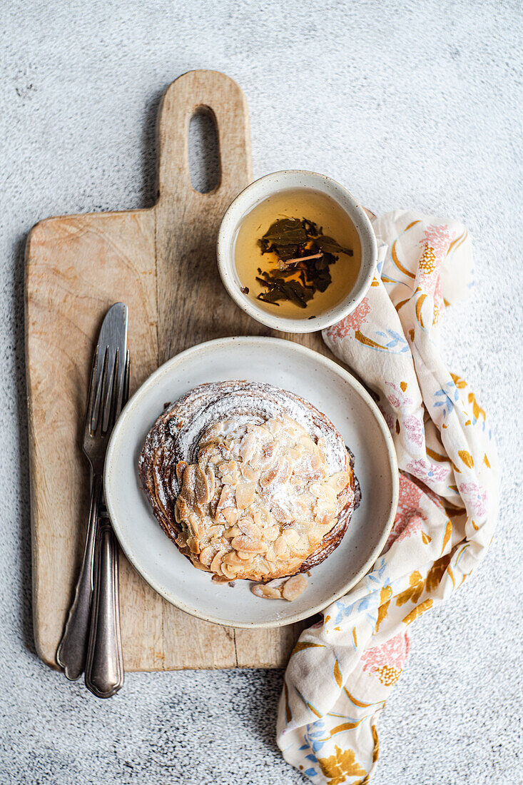 Draufsicht auf süßes Mandelgebäck auf Holzstäbchen zwischen Gabel und Messer und Serviette mit Tasse grünem Tee vor grauem Hintergrund