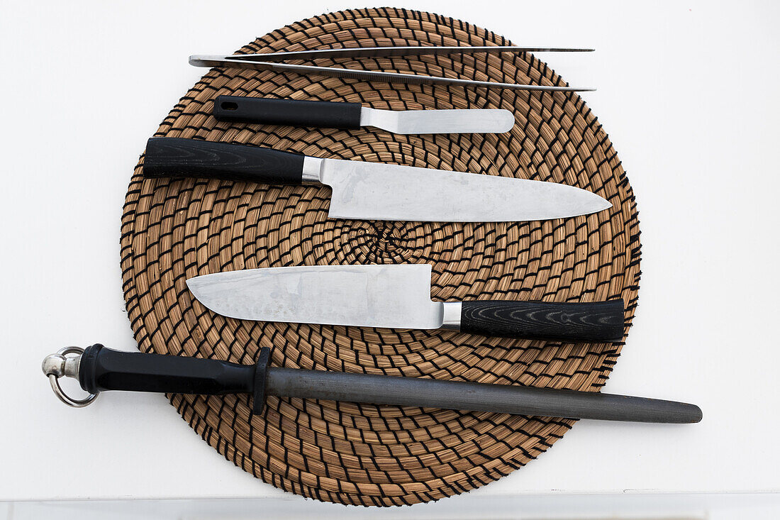 Top view of various types of knives comprising tongs bead cut chef utility and sharpening rod while placed on wicker placemat on kitchen table in daylight